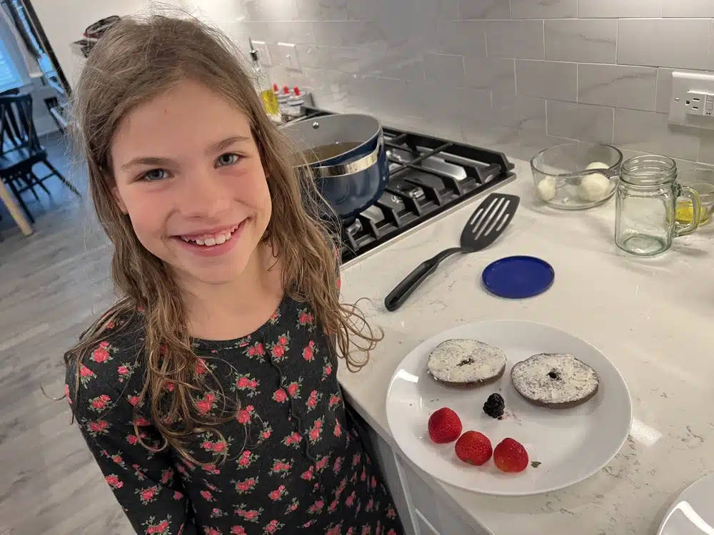 girl smiling and holding a plate with 2 homemade bagel halves and fruit arranged to make a smiley face